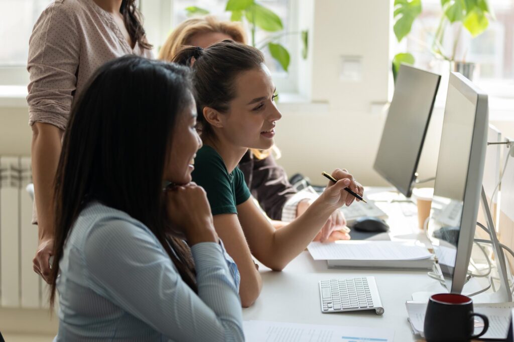 two women working on a desktop computer with colleagues in the background